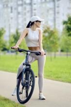A slender woman wearing activewear and a cap takes a break from cycling. She stands beside her bike