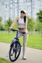 A woman sits on her bicycle in a vibrant park, dressed in a light sports outfit. She appears