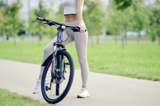 Slender female legs in athletic wear stand beside the mountain bike on a smooth pathway in a park.