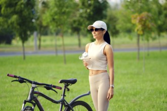 A woman in activewear holding a bottle with water stands beside her bicycle in a lush green park.