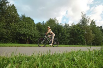 A woman in athletic wear rides her bicycle on a smooth path surrounded by lush green trees and