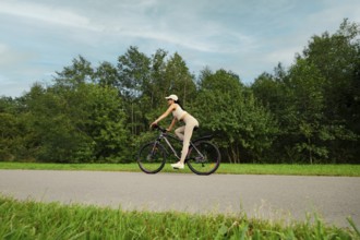 A woman rides her mountain bike on a smooth path in a lush green landscape. Tall trees line both