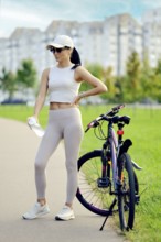 A woman in athletic wear stands on a path in a green park next to her bike, holding a water bottle.