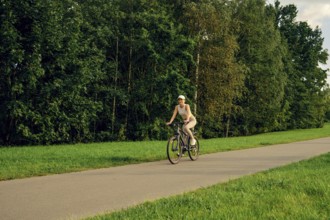 A woman rides a bicycle along a paved path surrounded by tall trees and vibrant greenery. The sunny