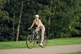 A slender woman is riding her bicycle on a smooth pathway bordered by lush greenery. She is dressed