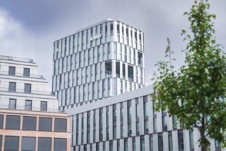 Modern high-rise building with glass façade under a cloudy sky, bordered by a tree, Munich, Germany