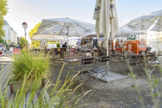 Cosy street café with empty tables and chairs under large parasols, surrounded by green plants,