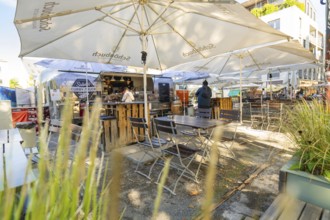 Outdoor area of a snack bar with tables and chairs under parasols, surrounded by plants,