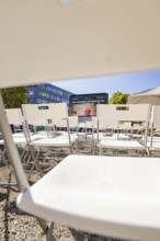 White chairs arranged in rows with a view of a screen and a building in the background, open air