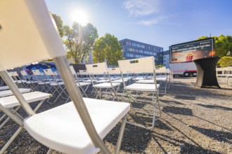 White chairs lined up outside in sunny weather in front of a building and a large screen, open air