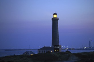 A lighthouse shines at dusk on the coast, surrounded by calm sea and blue sky, Skagen, Denmark