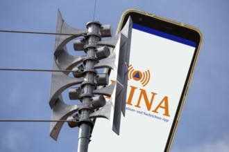 (Photomontage) Close-up of a siren, next to it the open warning app NINA on a smartphone