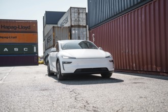 White car in the foreground of large, colourful freight containers in a port area, Tesla Model Y