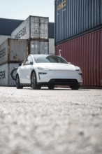 White car in a container port with different coloured freight containers, Tesla Model Y Juniper,