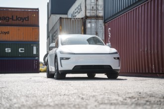 White car in a harbour environment next to stacked freight containers, Tesla Model Y Juniper,