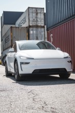 Modern white car in an industrial area surrounded by freight containers, Tesla Model Y Juniper,