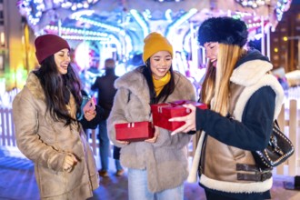 Three happy female friends exchanging christmas gifts in front of a carousel at a christmas market