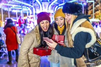 Three cheerful young women holding christmas gifts and using a smartphone in a christmas market