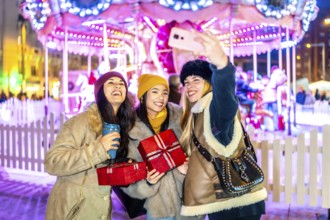 Three cheerful young women taking a selfie at a christmas market, holding gifts and warm drinks,