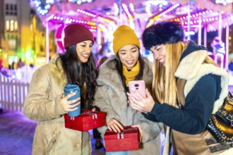 Three happy women using a smartphone and holding christmas gifts at night in a christmas market