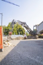 A large excavator and a construction worker on a building site with gravel and a lorry. Blue sky