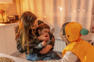 Mom applying halloween makeup on her son's face while his brother, dressed in a pumpkin costume,