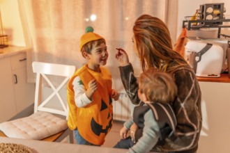 Mother applying halloween makeup to her son wearing a pumpkin costume while holding her baby in the