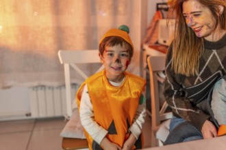 Happy child wearing pumpkin costume smiling and looking away while sitting next to his mother