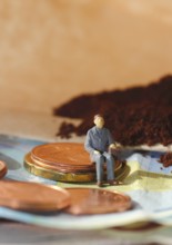 Miniature figure of a man sitting on coins and banknotes, ground coffee in a filter bag in the