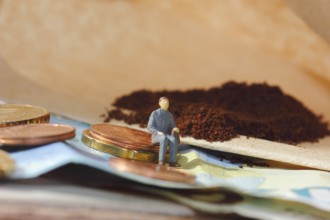 Miniature figure of a man sitting on coins and banknotes, ground coffee in a filter bag in the