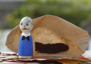 Male wooden cone figure with coins and banknote in front of filter bag with coffee North