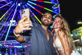 Young couple smiling and taking a selfie with smartphone in front of a colorful ferris wheel at an