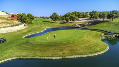 Two golfers are putting on a green surrounded by water, with lush fairways and trees in the