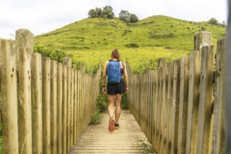 Female hiker with backpack walking on wooden footbridge leading towards a green hill in zumaia,