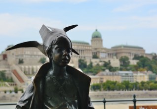 Bronze statue of a little princess wearing a bird shaped hat, overlooking the majestic buda castle