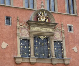 Architectural detail showcasing an ornate window with a coat of arms, latin inscription, and