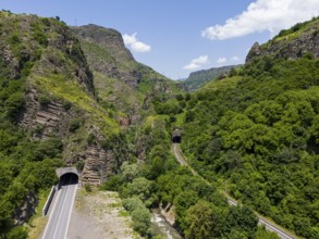 Road tunnel and railway tunnel through forested mountain landscape under blue sky, aerial view,