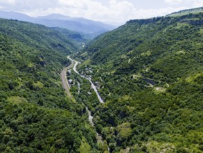 A green valley with dense vegetation, a road and a river between mountain ranges under a cloudy