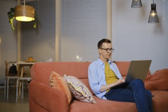 A man sits comfortably on a sofa and chats online on his laptop during evening hours. He