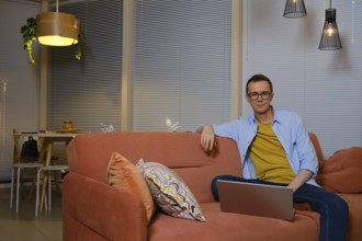 A man sits comfortably on a coral sofa, focused on his laptop in a cozy living room decorated with