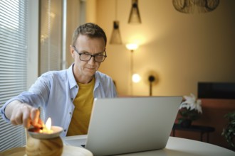 A middle-aged man lights small candle with lighter while working on his laptop at a round table in