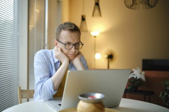 A middle-aged man with glasses watching series on his laptop while seated at a round table in a