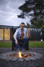 A man sits on a black chair, attentively tending to a fire pit surrounded by stones. The evening