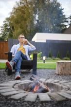 A middle-aged man sits in an outdoor chair, sipping a drink while enjoying the warmth of a fire pit