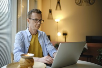 A middle-aged man sits at a table and typing letter on his laptop in a living room. Soft lighting