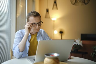 A focused man with glasses is working on his laptop with financial documents at home in the evening