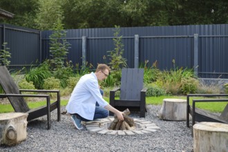 A middle-aged man dressed in a light blue shirt arranges firewood at a circular stone fire pit