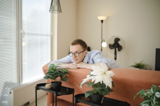 A middle-aged man leans on a colourful couch, gazing thoughtfully at potted indoor plants nearby.