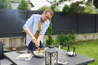 A man sprinkles fresh vegetables with salt in the backyard. Smoke from a barbecue can be seen in
