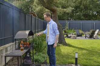 A man stands beside a barbecue grill, actively tending to the flames in a vibrant backyard. The